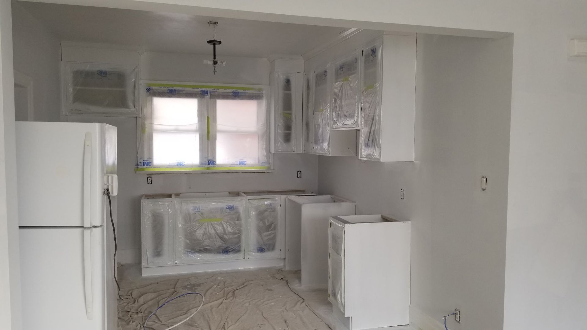 Kitchen with white cabinets covered in plastic for painting. A refrigerator is on the left.