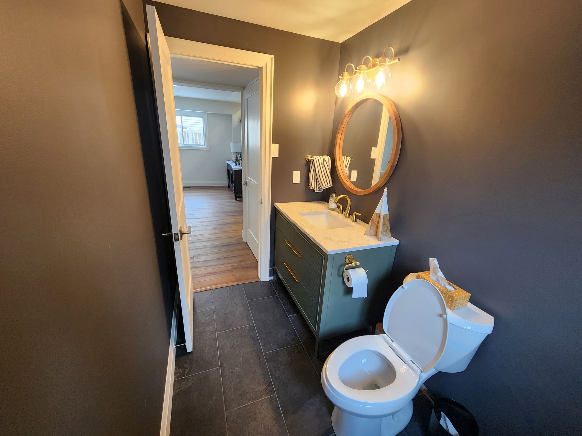 Bathroom with dark gray walls, green vanity, oval mirror, toilet, and open doorway to another room.