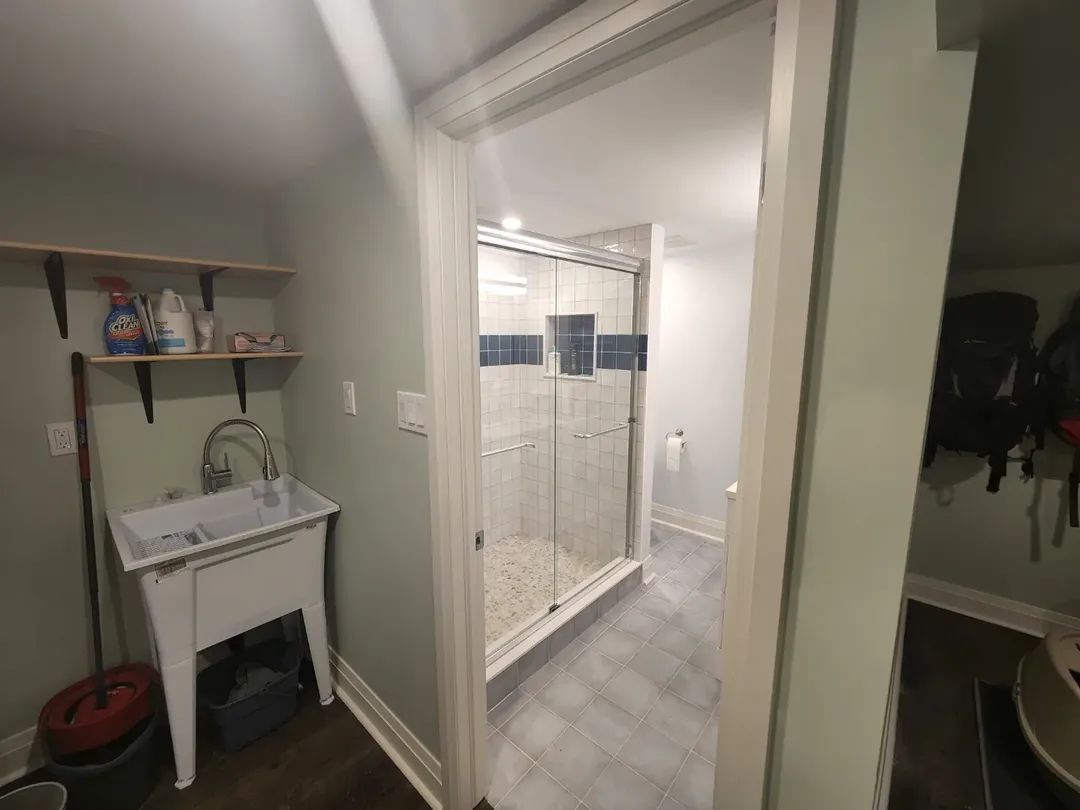 A utility room with a laundry sink, shelf, and a doorway leading to a bathroom with a shower.
