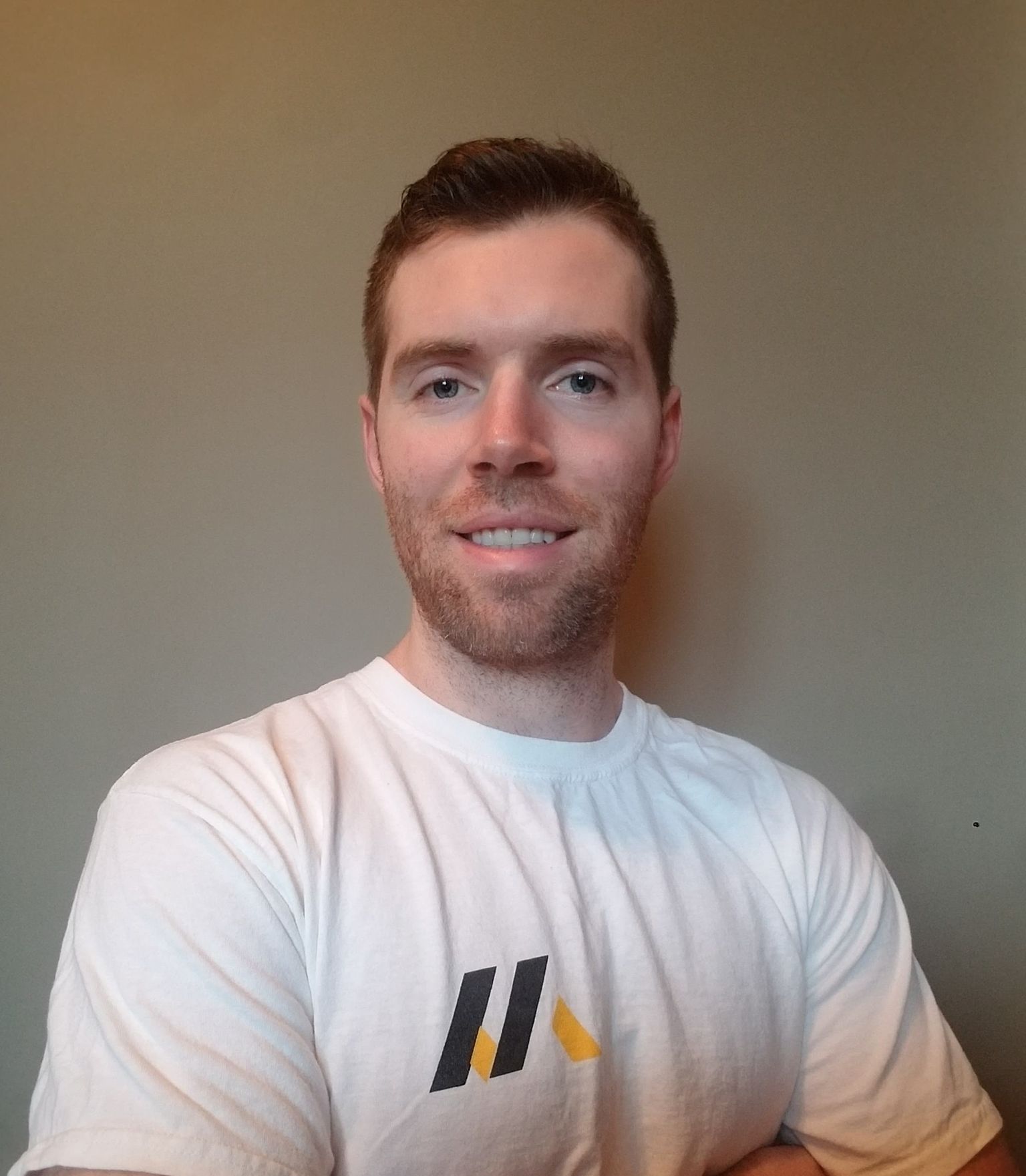 Man in white shirt with logo smiles in front of a neutral wall.