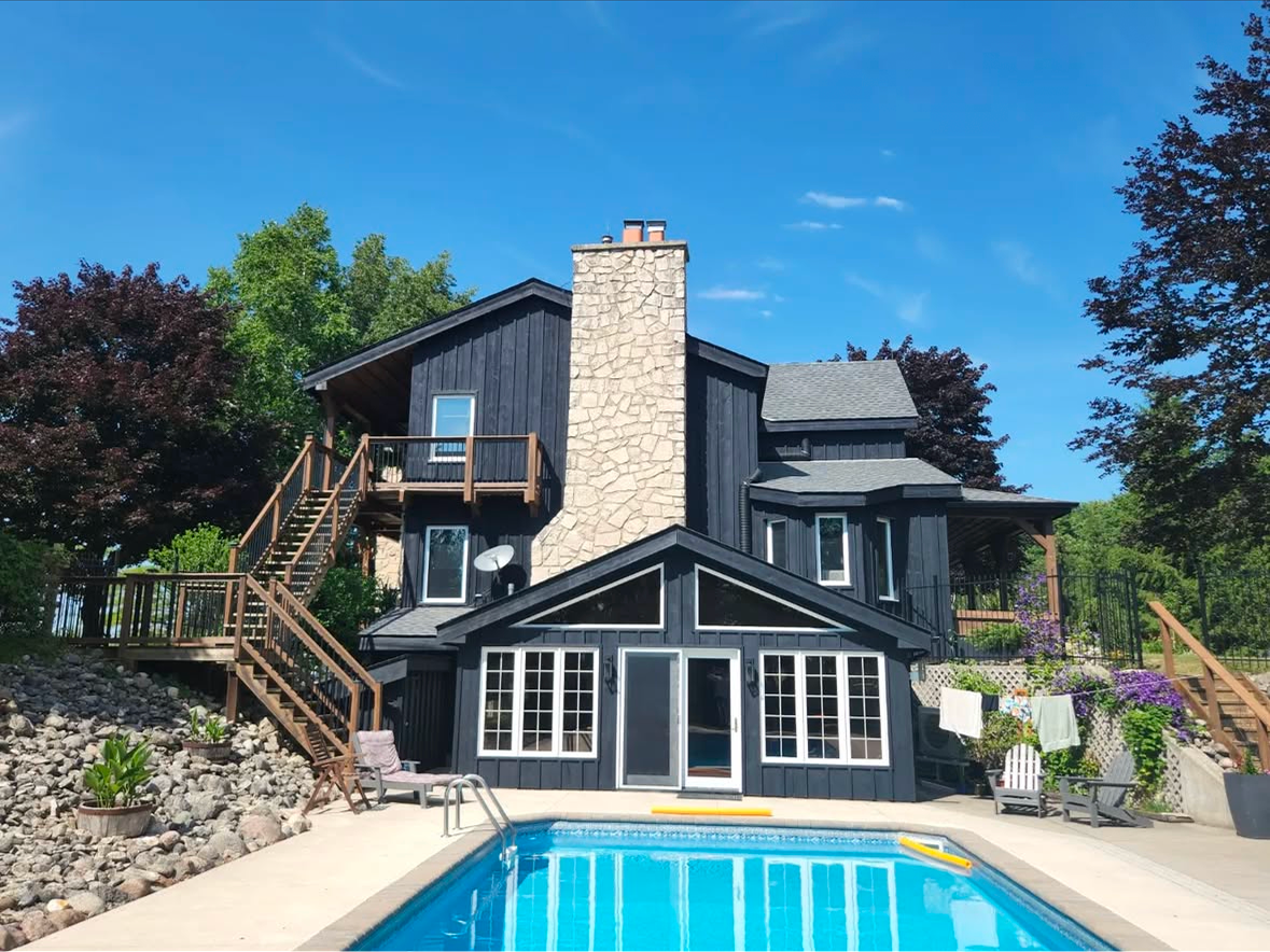 Black house with a stone chimney, pool, and wooden stairs against a blue sky.