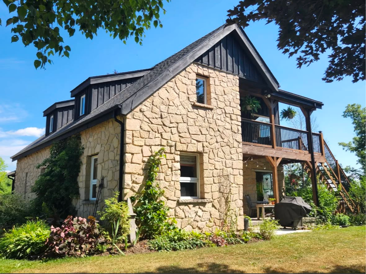 Stone house with black roof and balcony, surrounded by greenery.