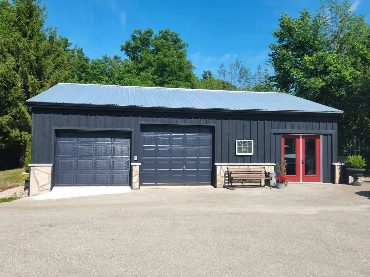 Dark blue garage with metal roof, two bays, red doors, and a bench; outdoors, trees in background.