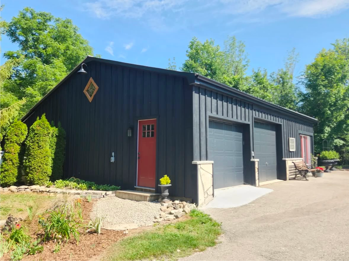 Black building with red door and garage doors; set in a yard with trees and blue sky.
