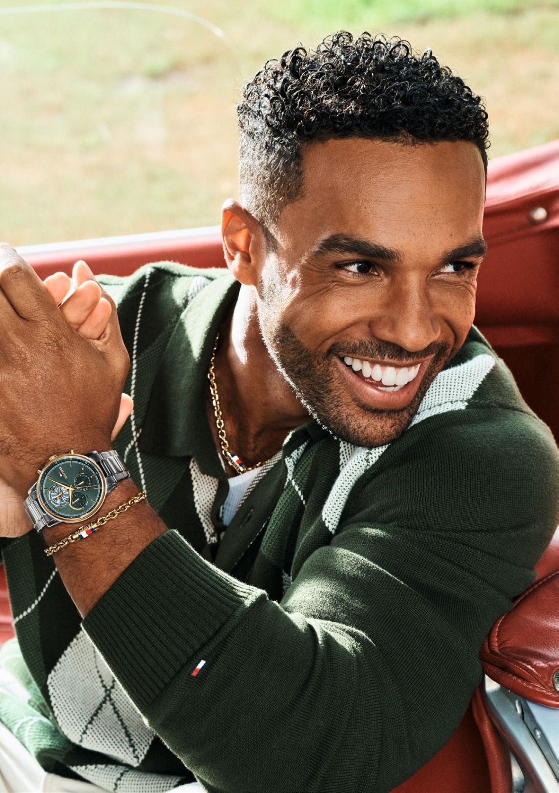 Un uomo sorridente, con indosso un maglione verde a rombi e un orologio, è seduto in una decappottabile rossa.