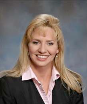 Woman with blonde hair wearing a pink collared shirt and black blazer smiles in front of a blue background.