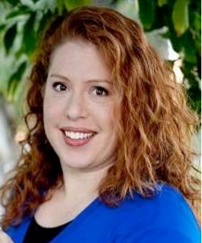 Woman with reddish-brown curly hair smiles, wearing a blue top against a blurred outdoor background.