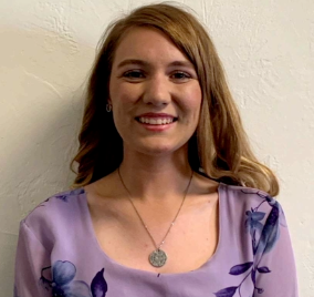 Woman with light brown hair, smiling, wearing a lavender floral shirt and a silver pendant necklace, against a cream wall.