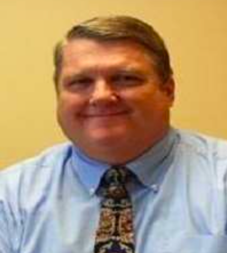 Man in light blue shirt and patterned tie smiles.