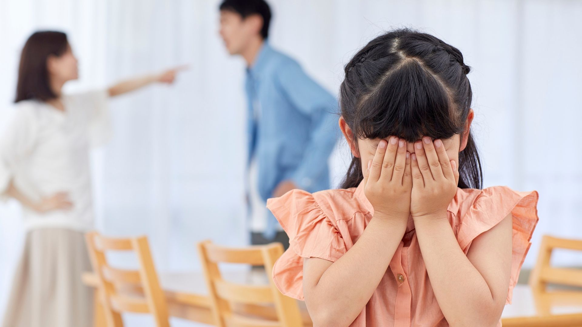 Child covering face, parents arguing in background.