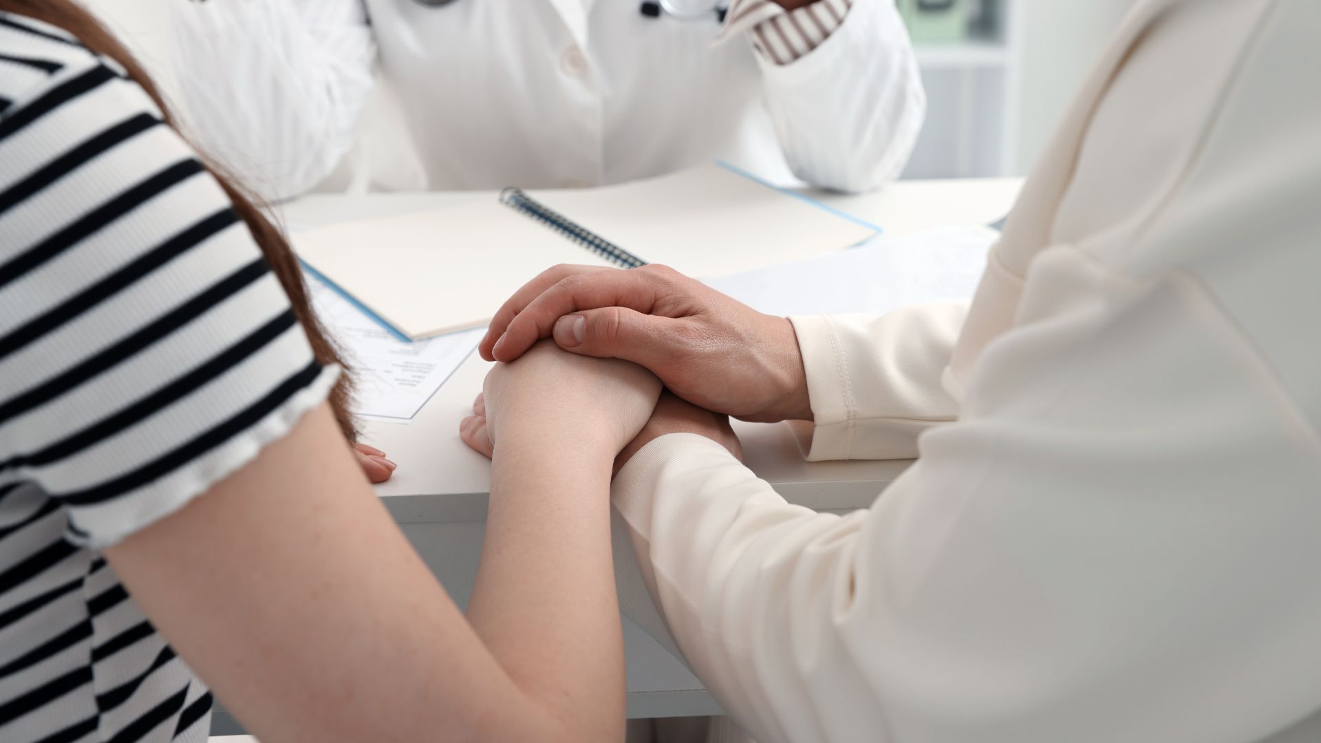 Doctor holding patient's hand for support at a white desk in a medical office.