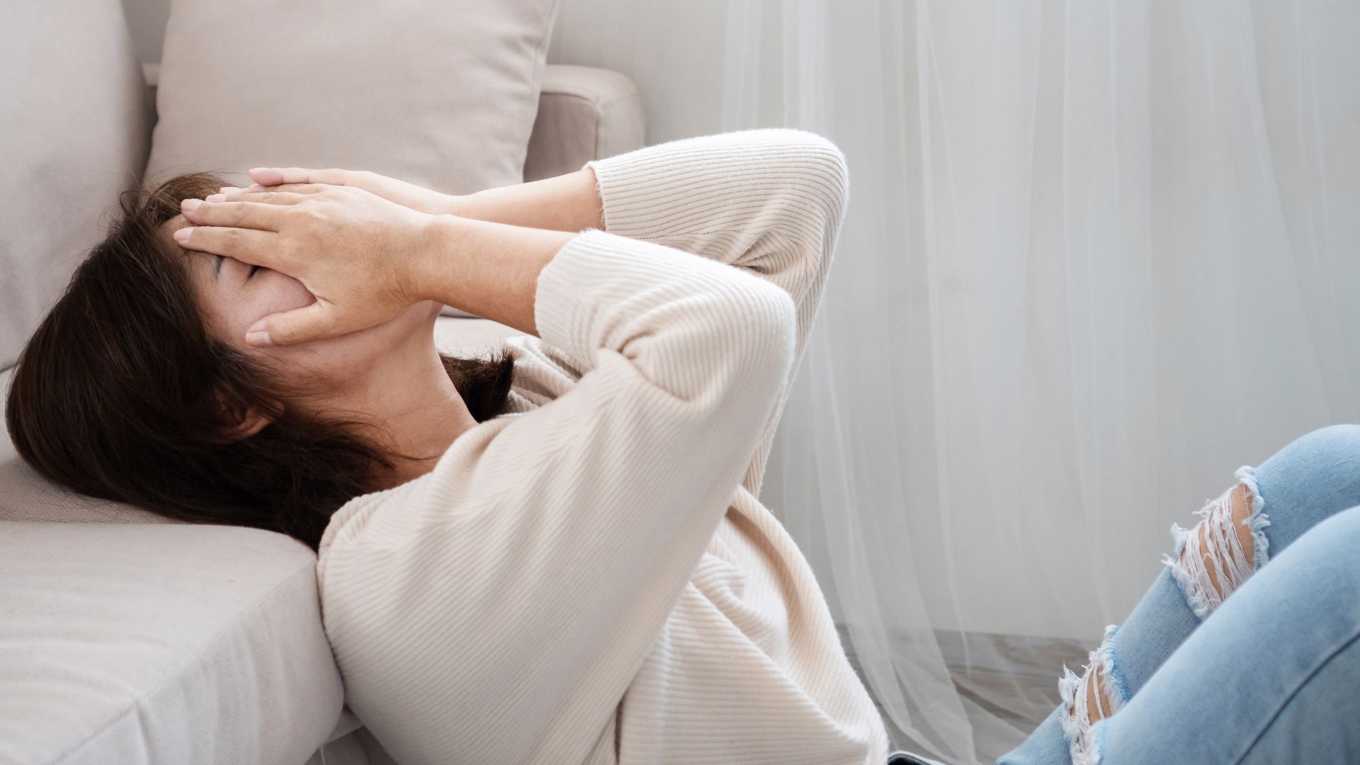 Woman sitting against couch, face covered by hands, appearing distressed.