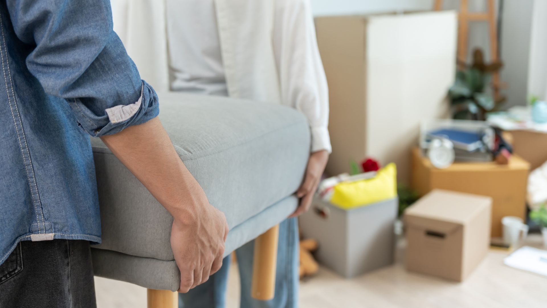 Two people carrying a gray upholstered ottoman, moving boxes in the background.