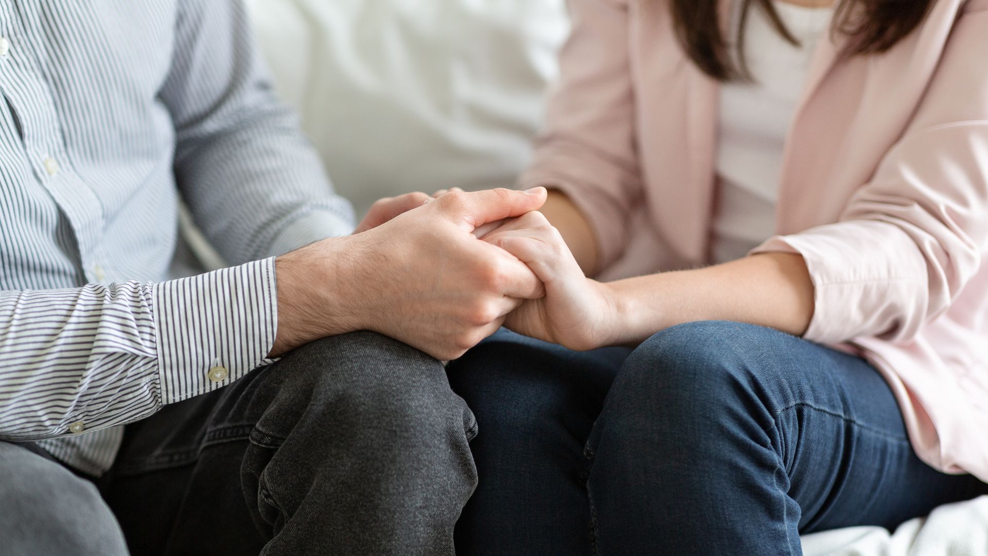 Couple holding hands on a couch; man's striped shirt, woman's pink jacket and jeans, emotional support.