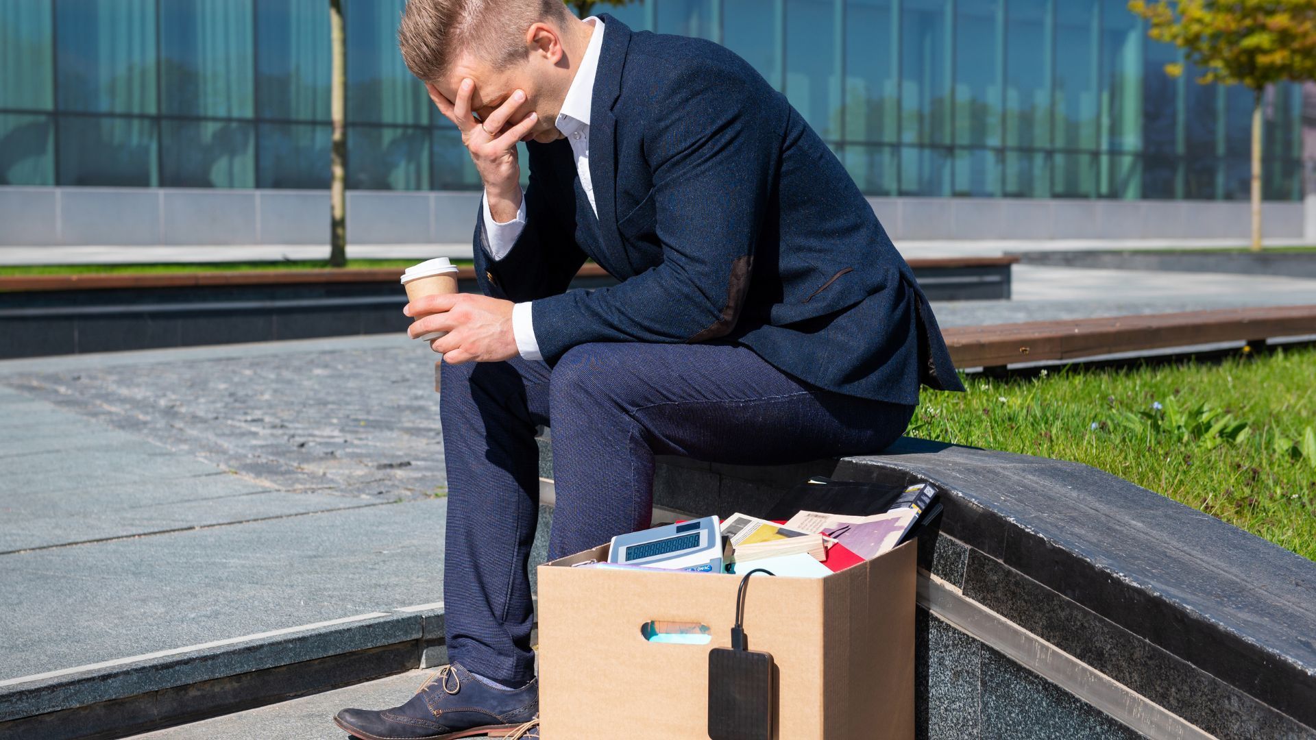 Man in suit sits outside, head in hand, near box of belongings; appears distressed.