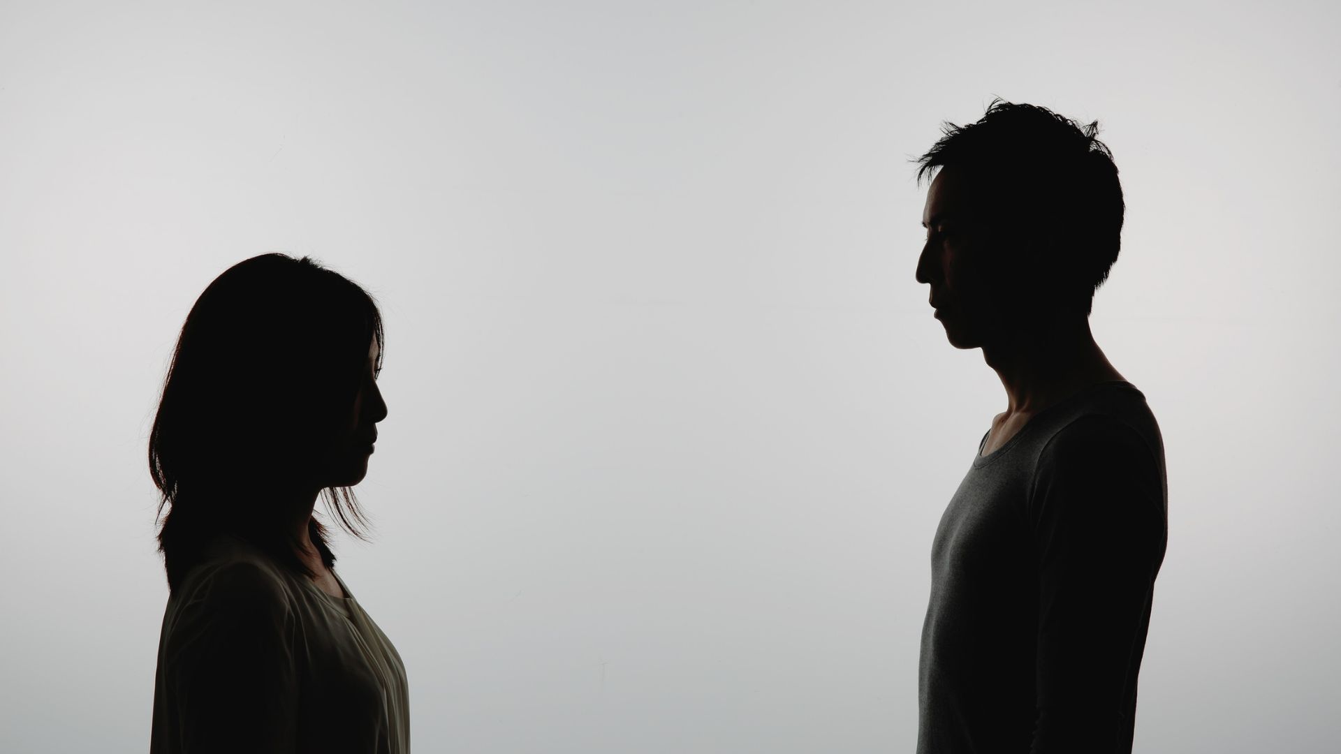 Woman sitting on a white sofa looking thoughtful while a man reads a newspaper behind her.