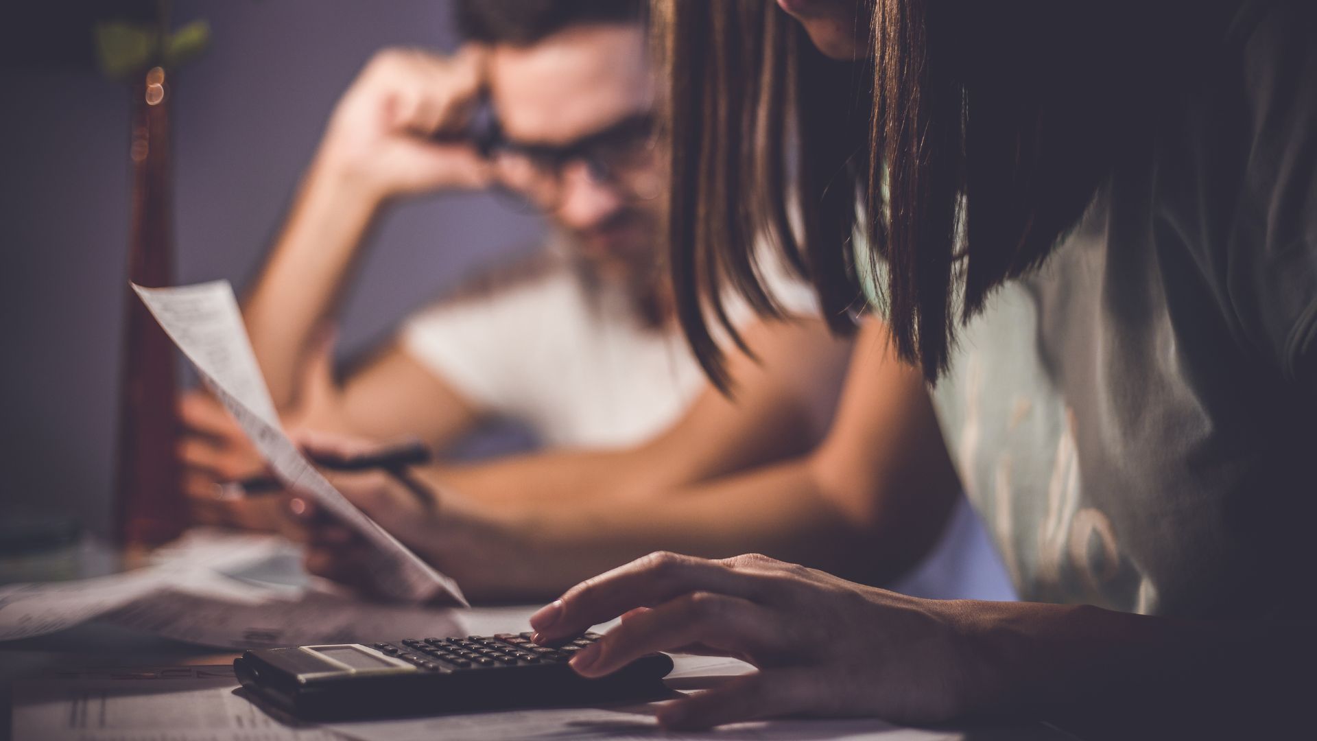 Woman and man at table with bills, looking stressed, using calculator and laptop.