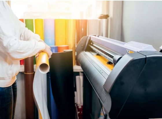 Persona con un rollo de vinilo amarillo cerca de una cortadora de vinilo y rollos de vinilo de colores. Taller.
