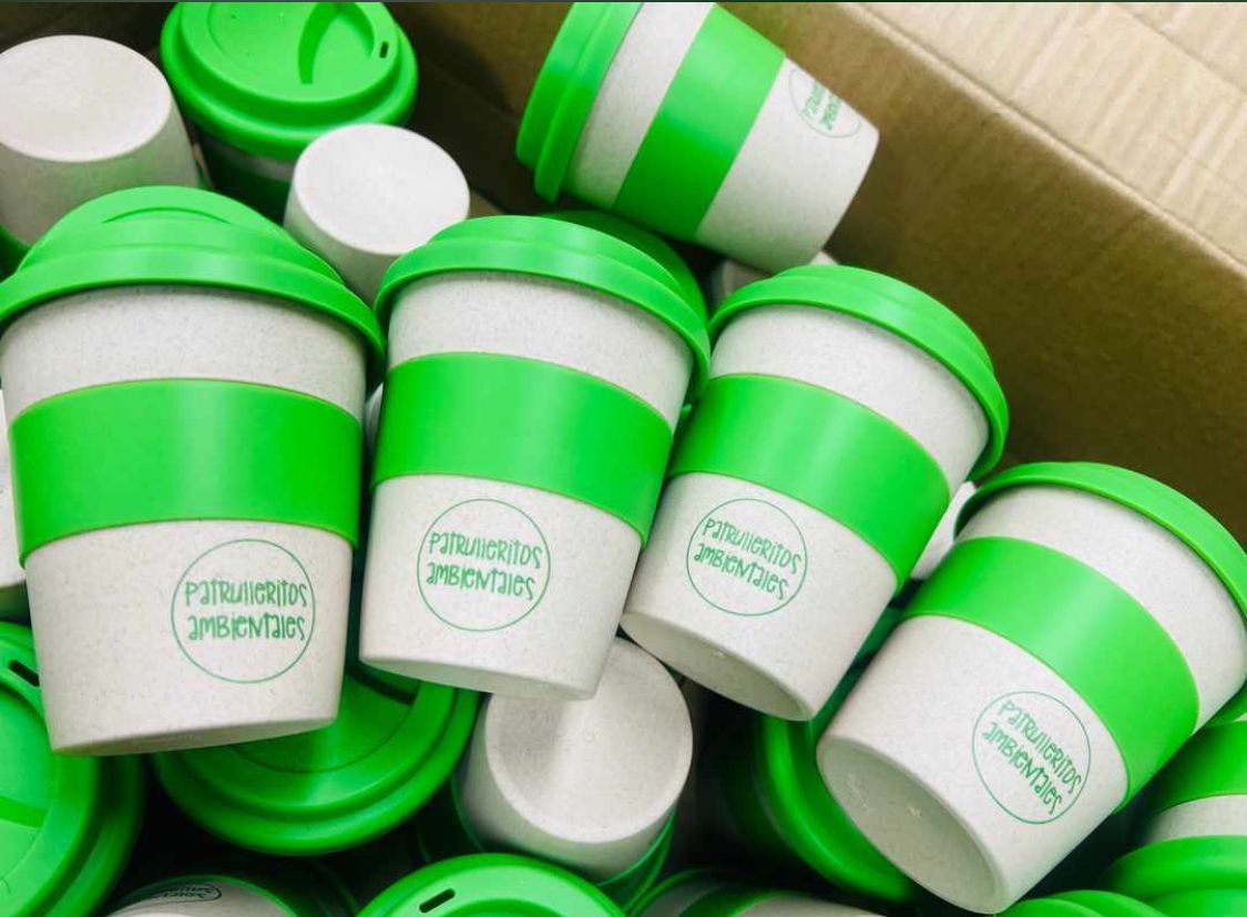 Vasos de café reutilizables verdes y blancos con tapa, apilados dentro de una caja de cartón.