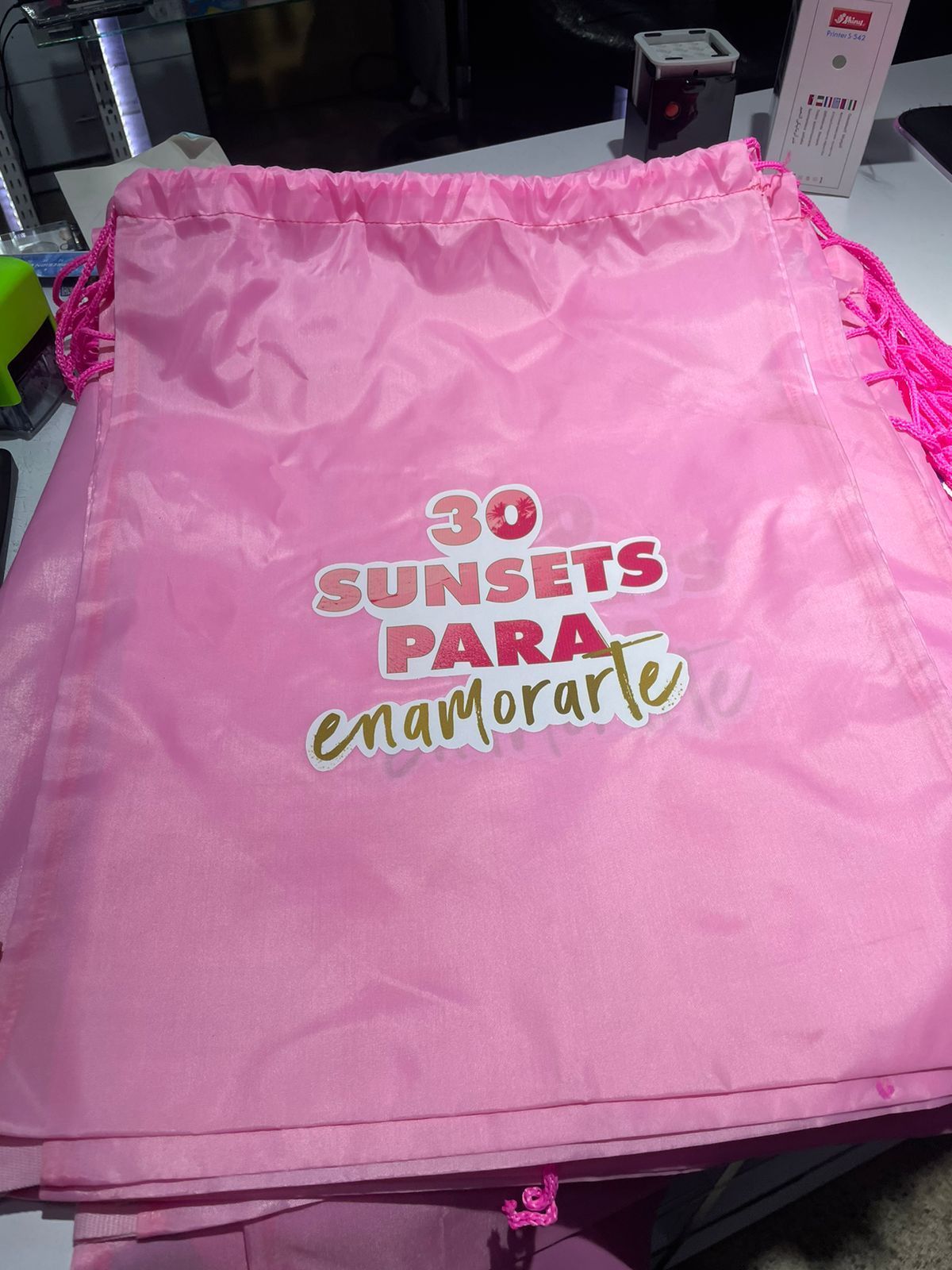 Bolsa de cordón rosa con texto “30 ATARDECERES PARA enamorarte” en rosa y dorado.