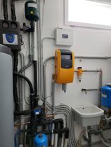 Locale tecnico con impianti idraulici e di riscaldamento, inclusi tubi, serbatoi, contatori e un'unità arancione a parete.