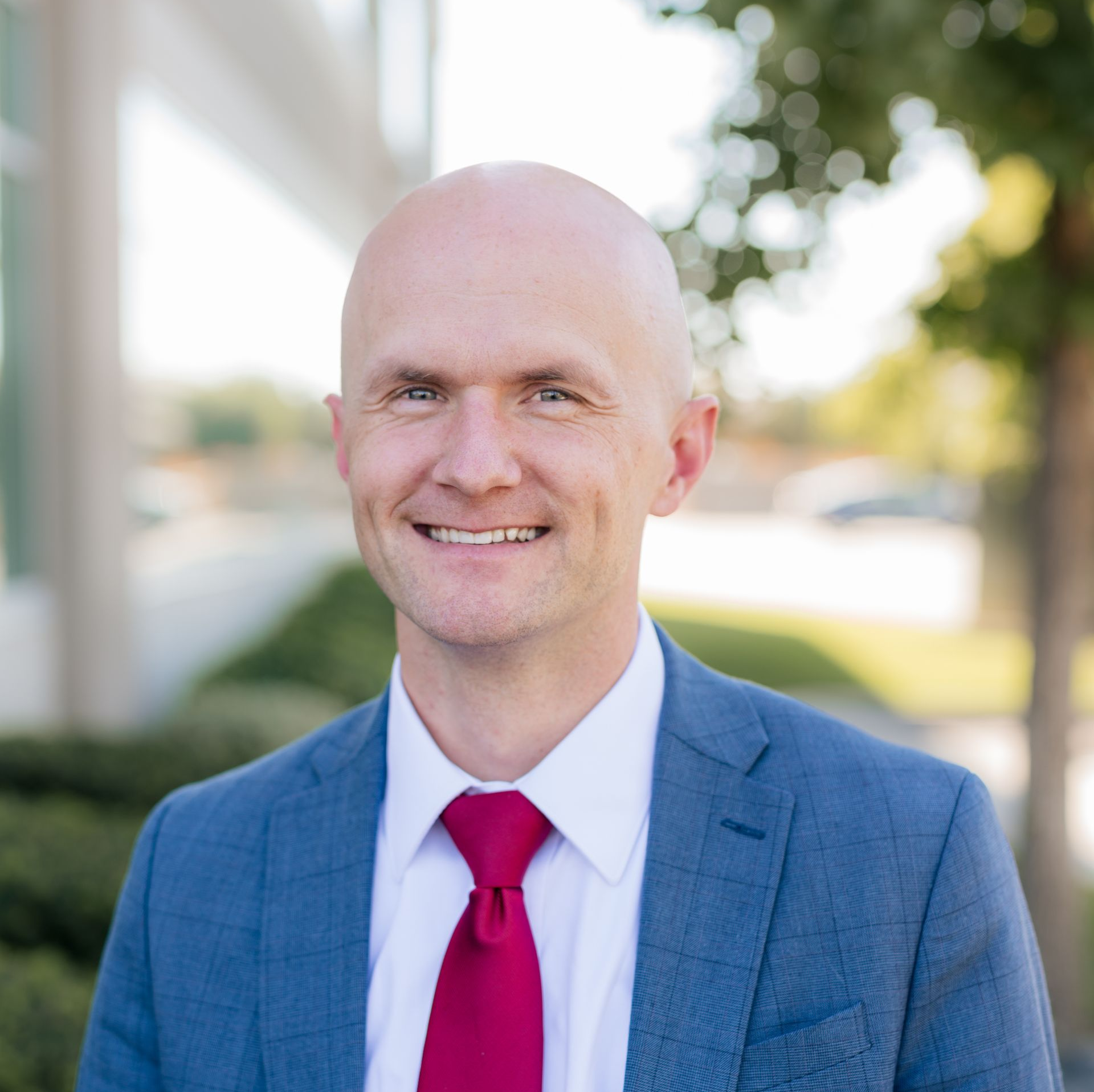 Bald man in a blue suit and red tie smiling outdoors near a building.