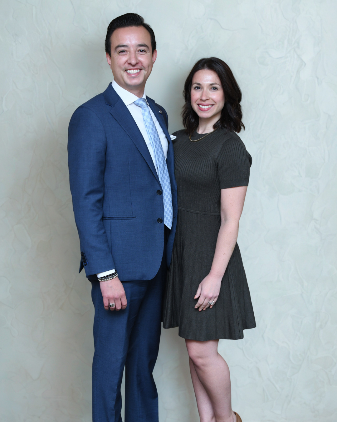 A person in a blue suit and tie stands beside a person in a dark dress, both smiling in front of a neutral background.