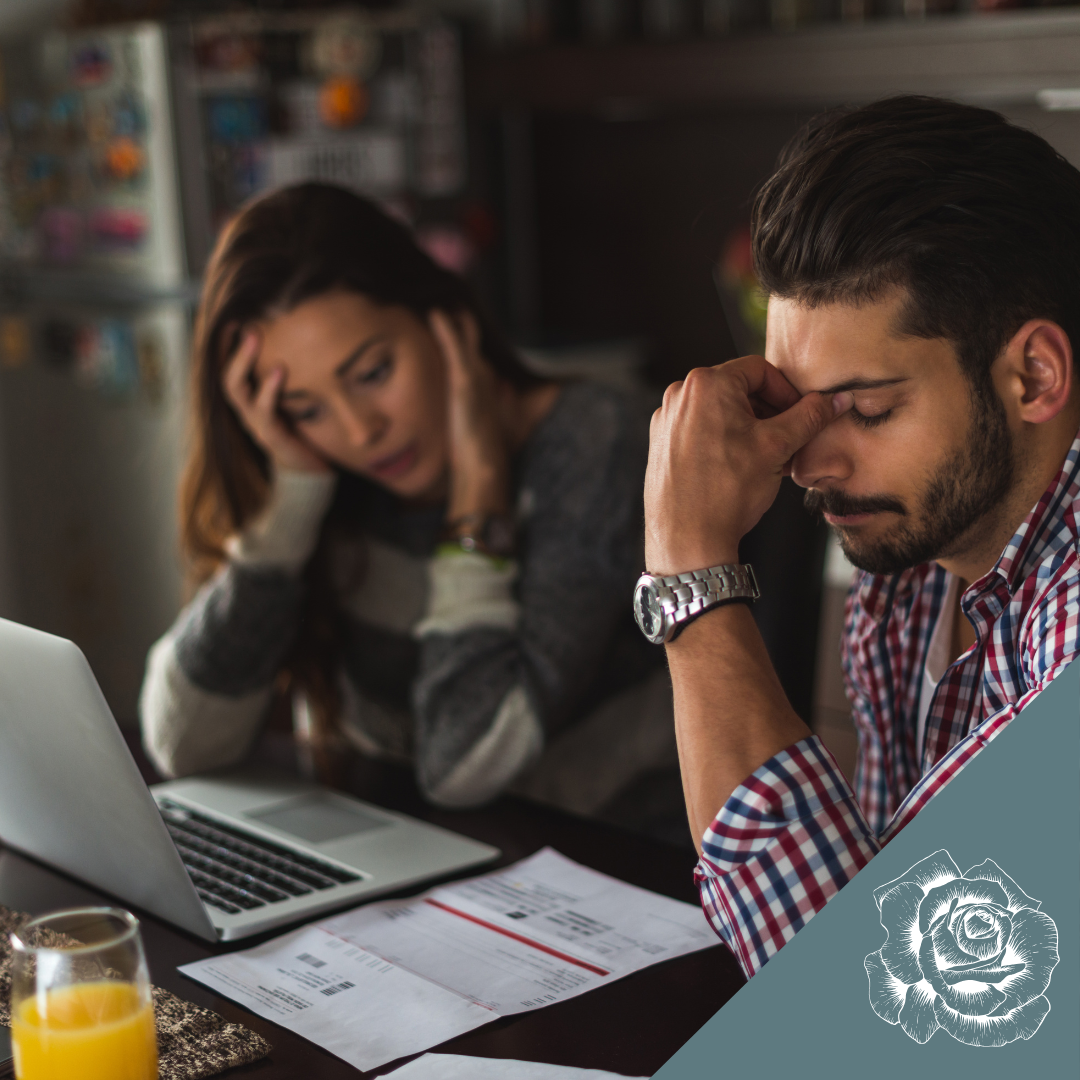 A stressed couple sits at a table with a laptop and documents, both holding their heads with expressions of frustration.