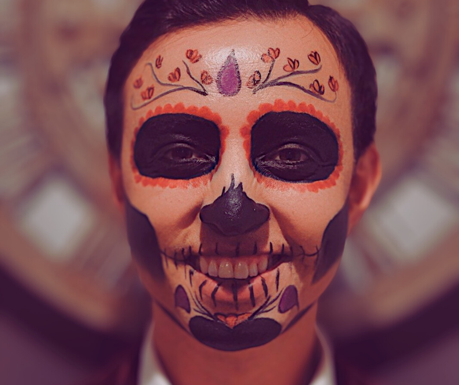 A person with Sugar Skull face paint, featuring black eye circles, a painted nose, and floral designs on the forehead.