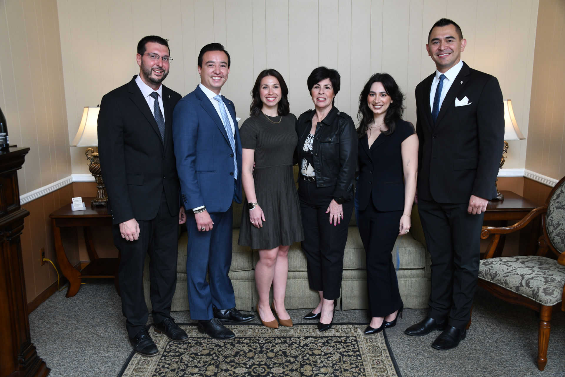 Six people in formal business attire pose standing in a row in a room with a sofa, lamps, and a patterned rug.