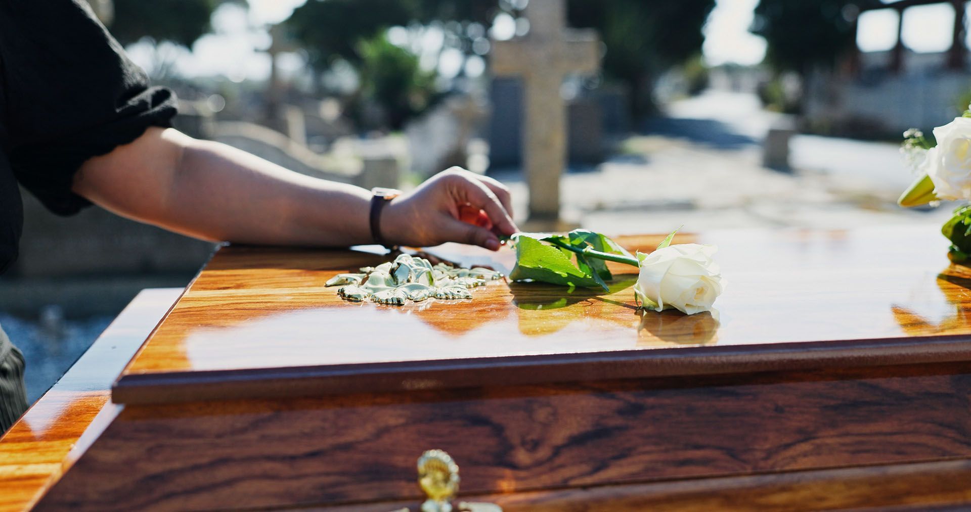 A person places white flowers and greenery on a wooden casket in a cemetery with a stone cross in the background.