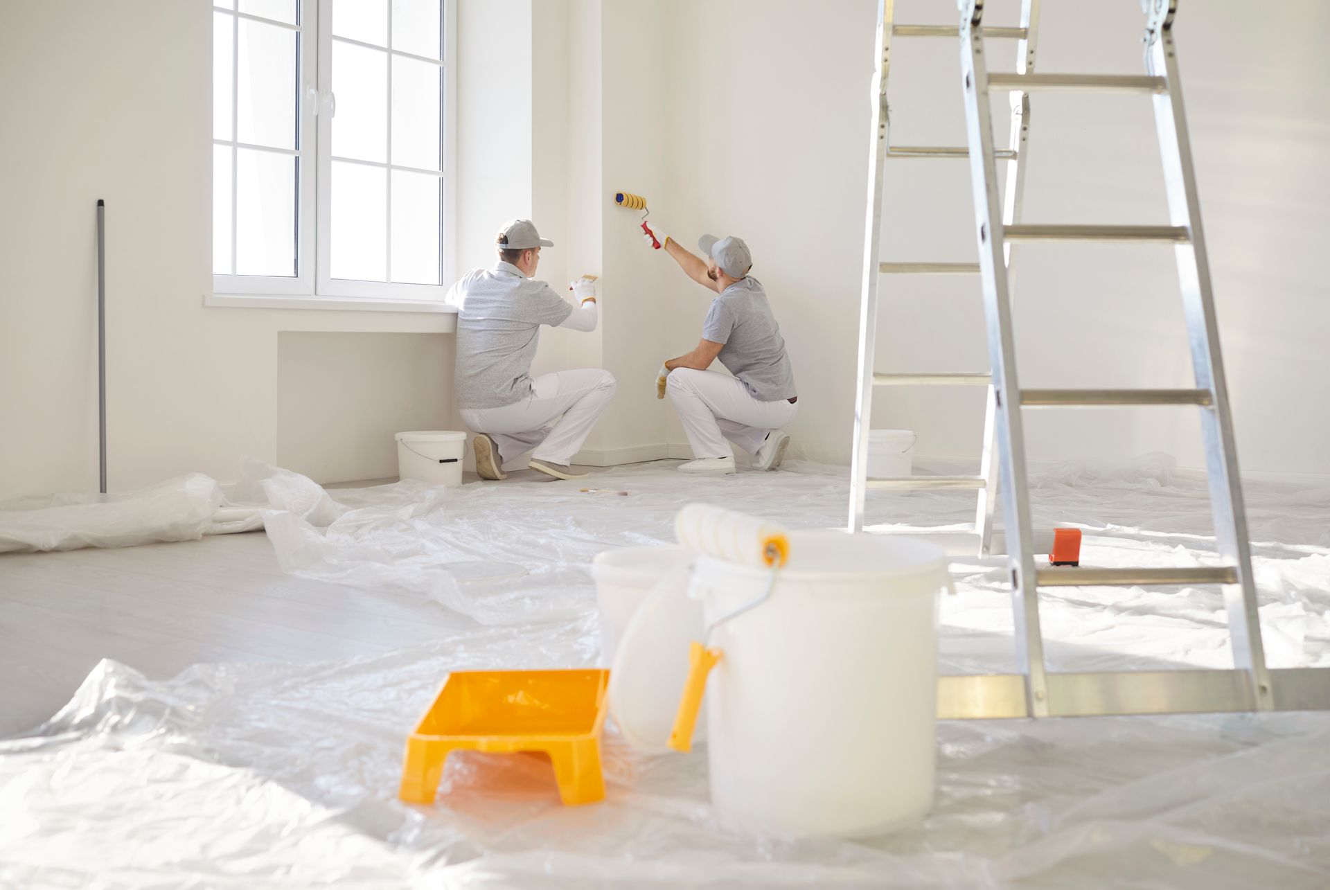 Two people painting a white wall in a room, with painting supplies and a ladder.