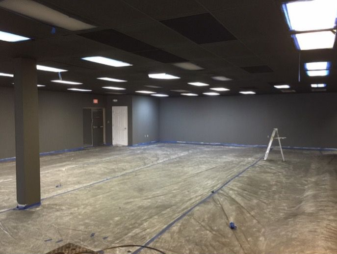 Empty room with gray walls, dark ceiling, concrete floor, and fluorescent lights. Tape lines the floor.