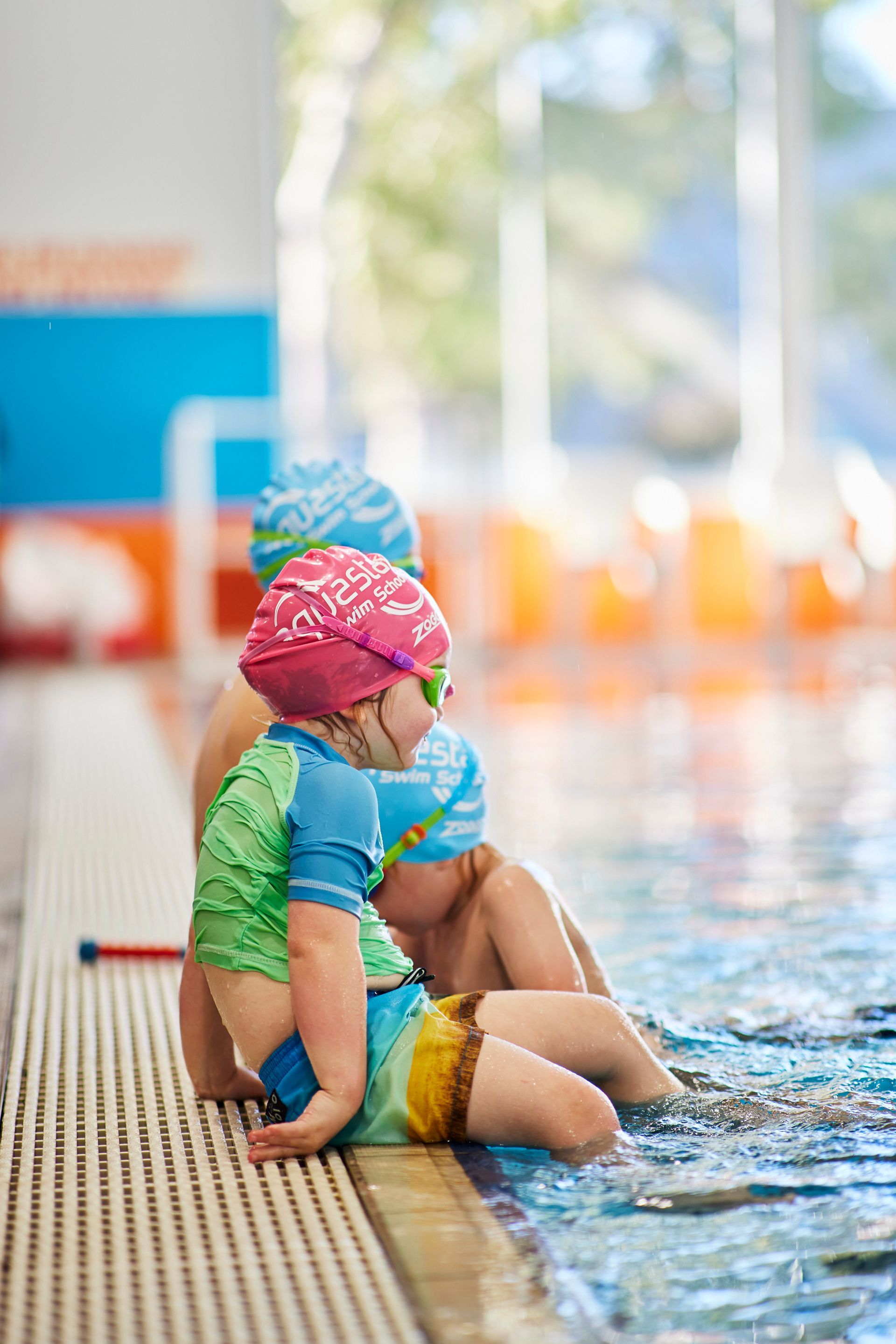 Children in swimsuits sit on pool edge, wearing caps and goggles.