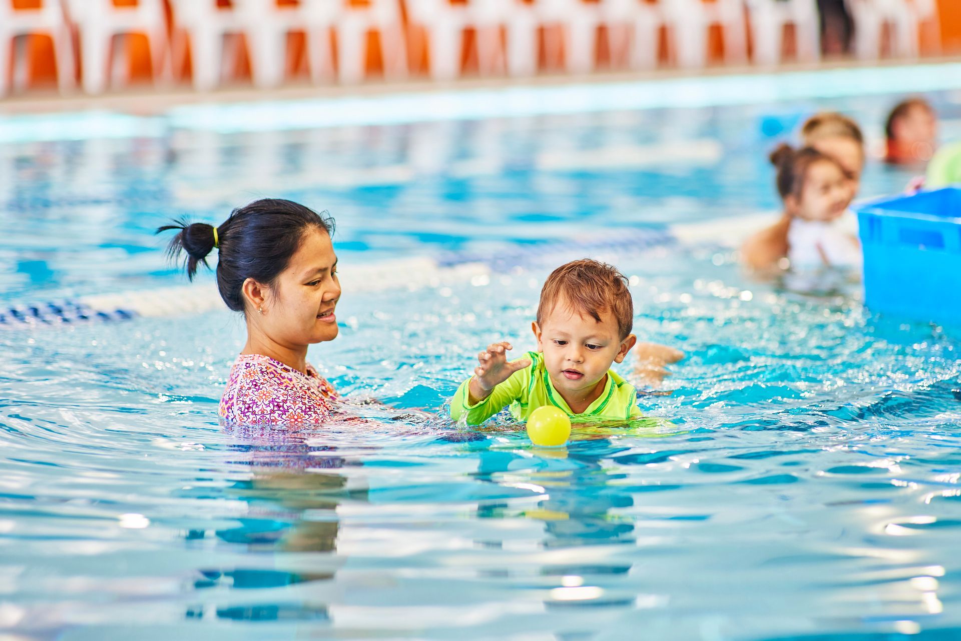 Instructor and child performing a swimming drill in a pool. Child in orange swim shorts, instructor in dark shirt.