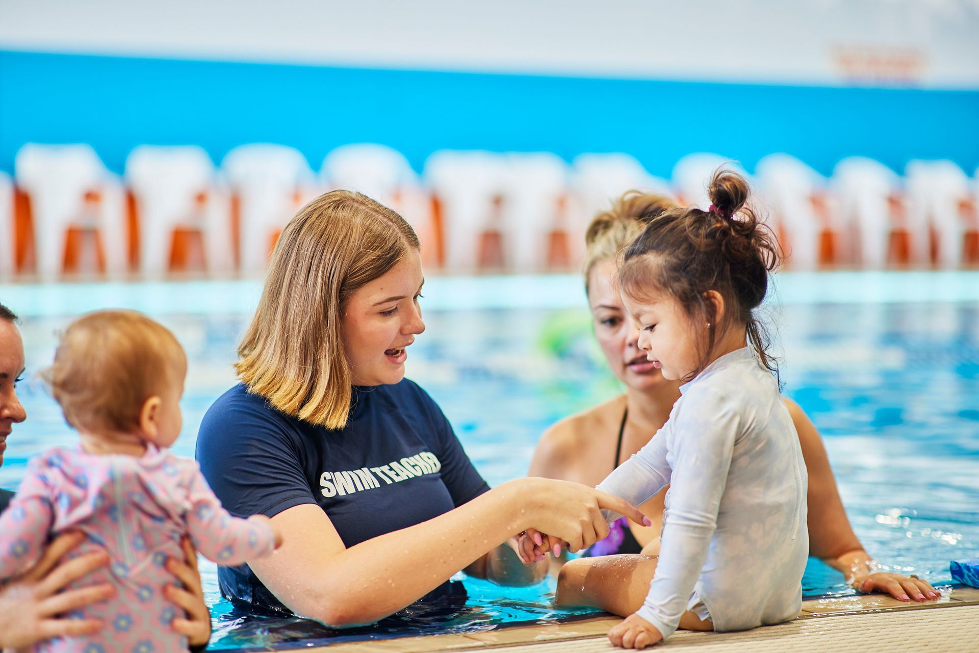 A swim instructor helps two toddlers in a pool, while their mothers watch.