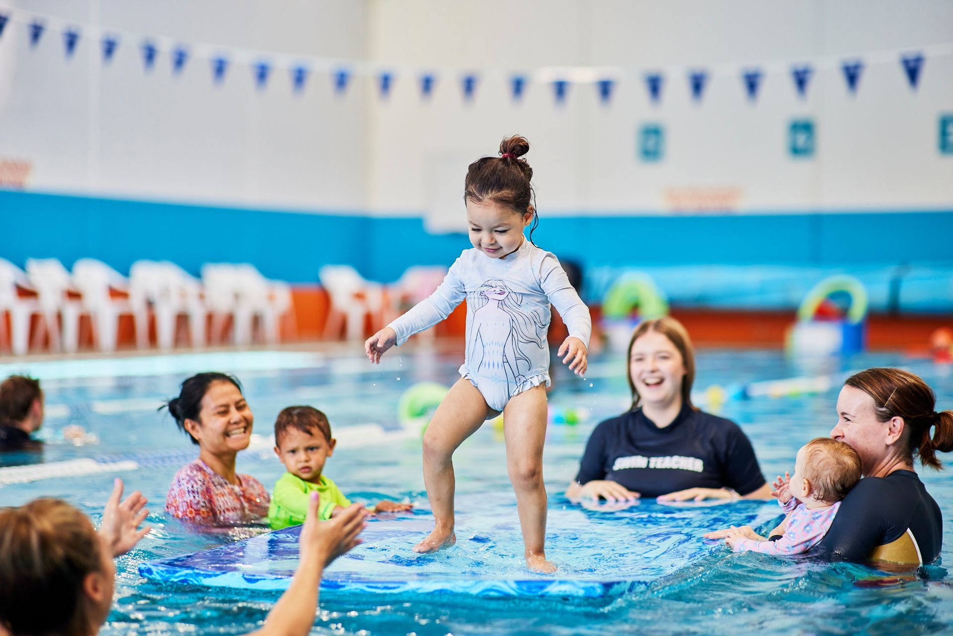Child wearing swimsuit stands on mat in pool, with adults, smiling, in a swimming pool.