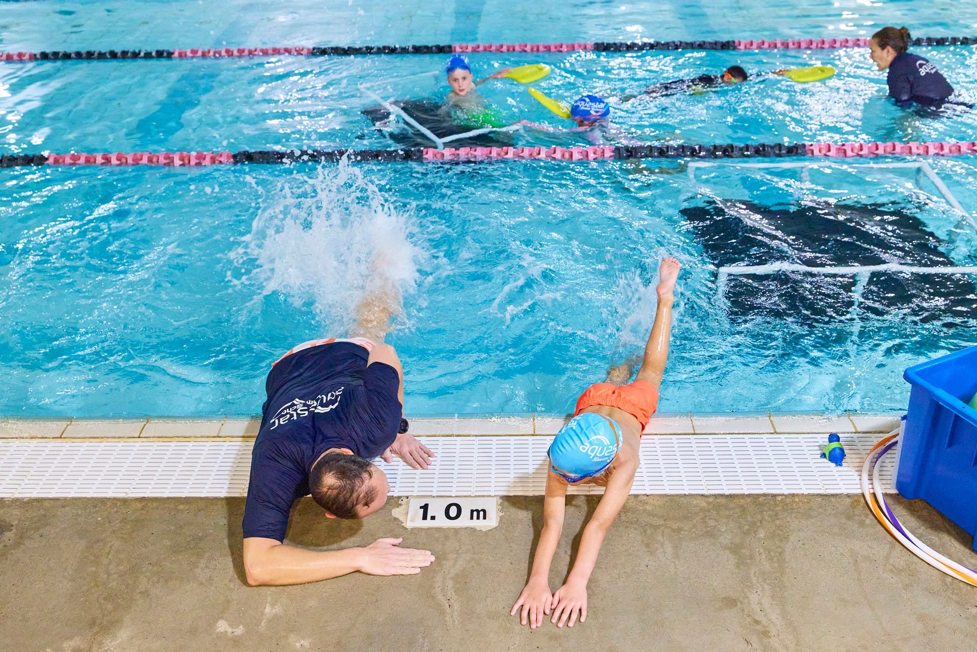Instructor and child performing a swimming drill in a pool. Child in orange swim shorts, instructor in dark shirt.