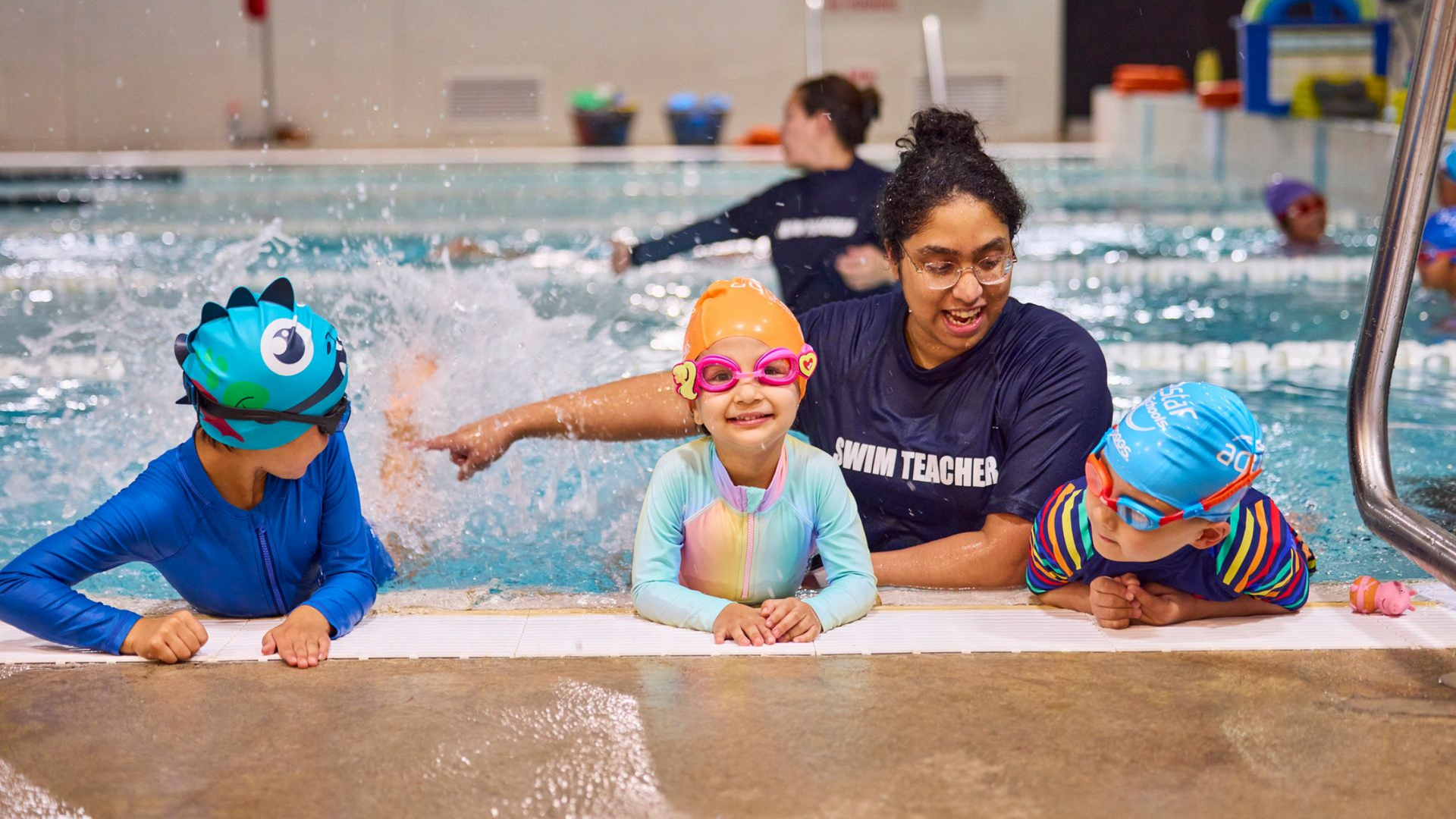 Swim instructor with three children in a pool, splashing water. Instructor points. All wear swim caps.