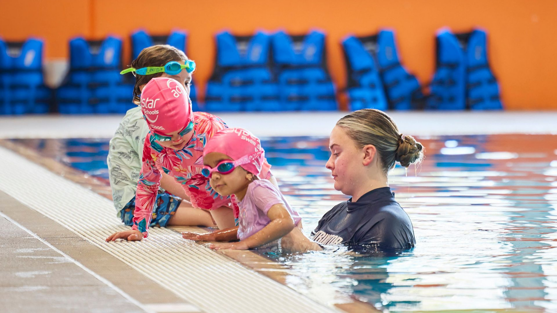 Children in swim gear, with instructor, entering a pool. Bright orange wall. Life vests.