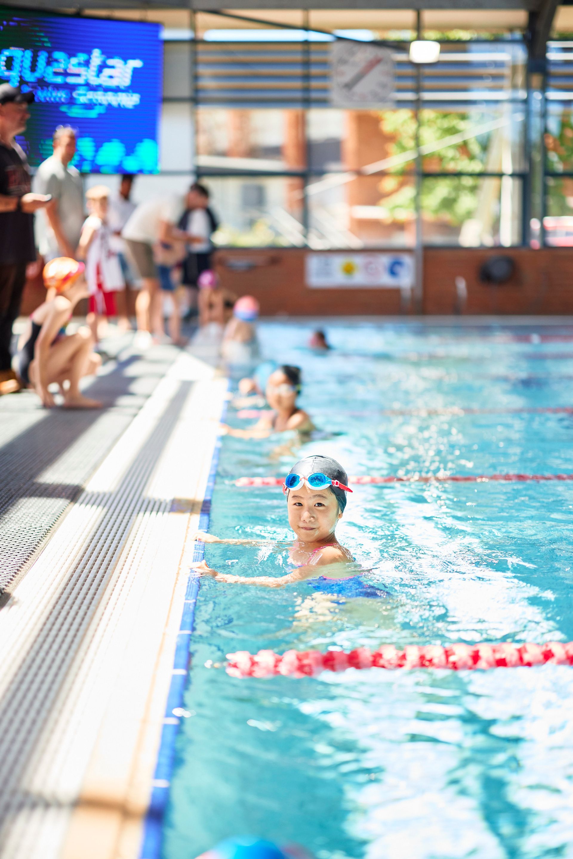 Young girl in blue swimsuit and goggles in a pool, looking at the camera. Other children and adults watch.