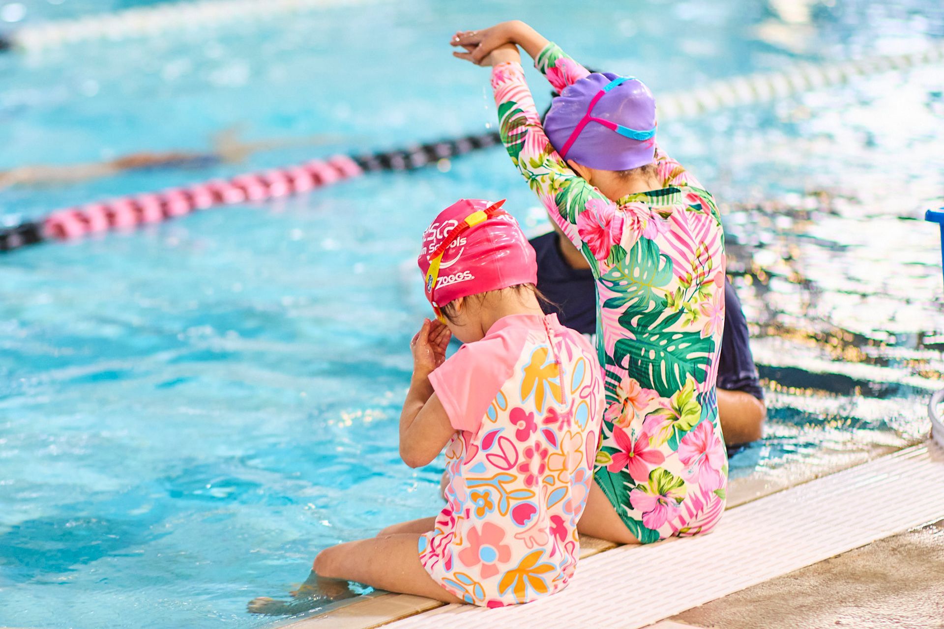 Two young girls in colorful swimwear sit by a pool, one adjusting goggles.