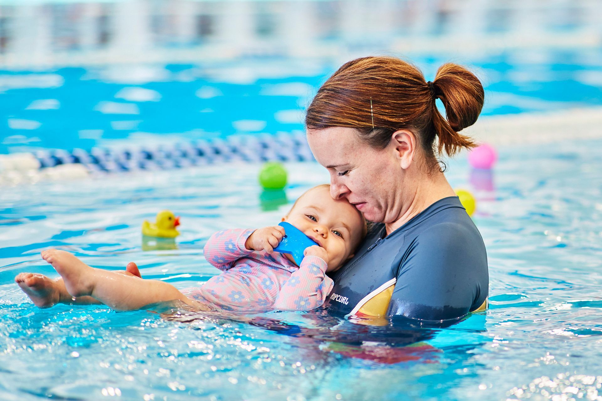 Woman in blue shirt in a pool, holding a baby wearing pink, both smiling.