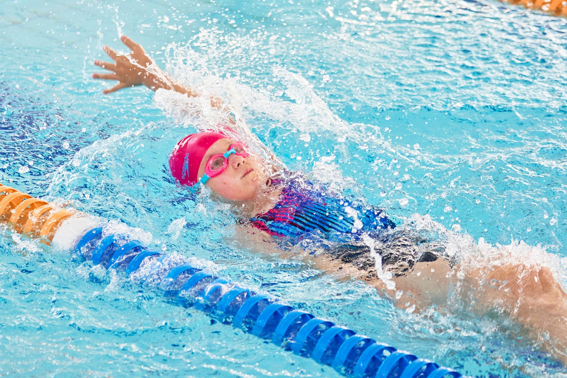 Young swimmer in a pink cap and goggles doing backstroke in a pool, water splashing.