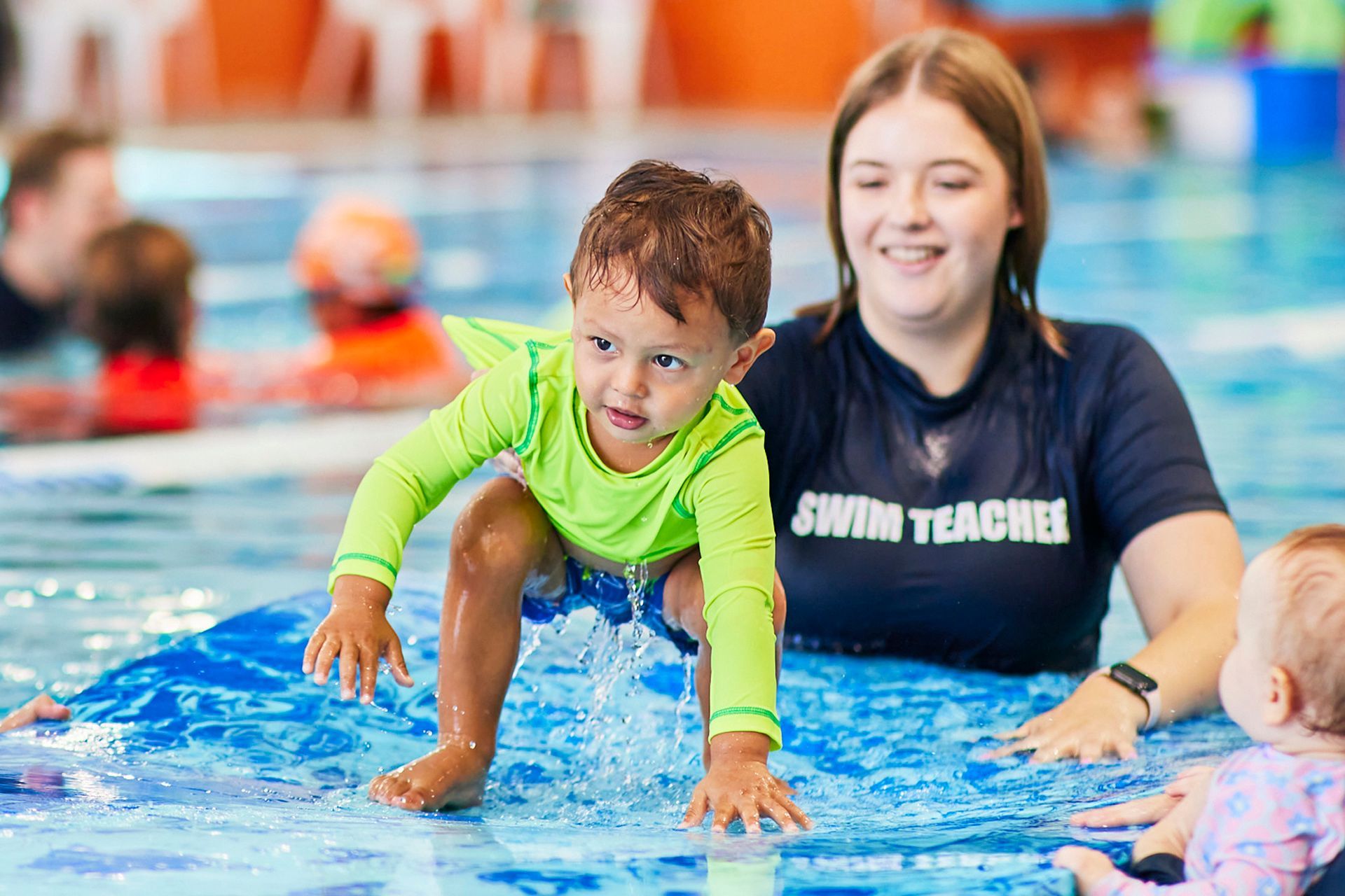 Young child in a green shirt practices swimming with a swim teacher in a pool; other children in the background.