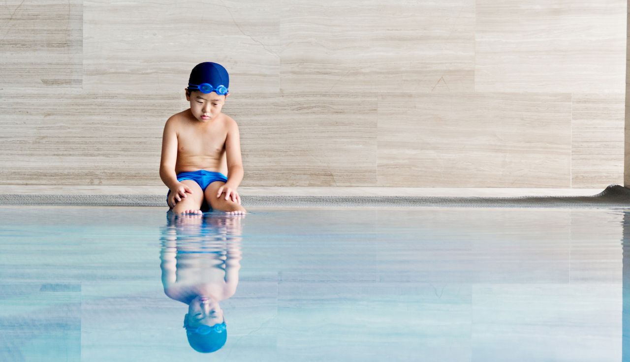 Young boy in swim trunks and cap, sadly sitting at edge of pool, reflection in water.