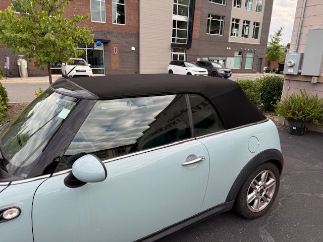Light blue Mini Cooper convertible parked outdoors. Black top, silver trim, brick building background.