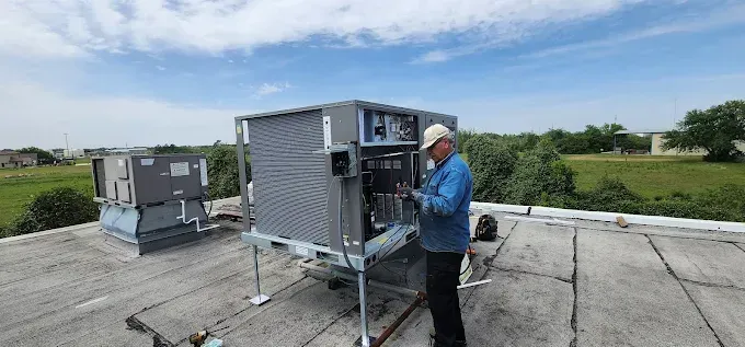 Technician inspecting rooftop HVAC unit on a concrete roof with another unit nearby under a blue sky