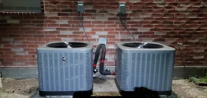 Two gray outdoor AC units on gravel beside a brick wall, connected by black hoses.