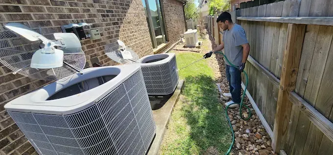 Man spraying weeds along a narrow side yard beside large air conditioner units and a wooden fence